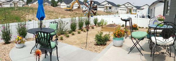 wide angle photo of yard with patio furniture, raised vegetable garden in back, shrubs along a white fence